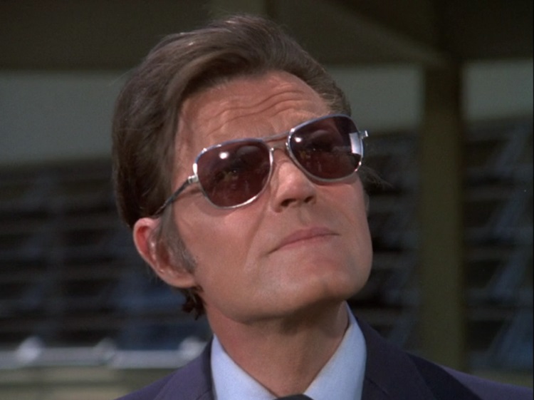 Jack Lord as Steve McGarret, a serious looking white man in his fifities with brown hair wearing silver framed sunglasses and a dark blue suit.