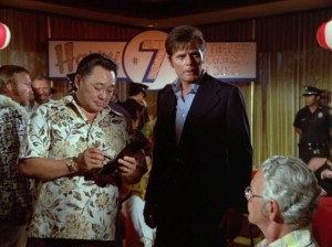 Chin, a Chinese man with black hair wearing a brown, beige, and white aloha shirt, and Steve, a white man with brown hair wearing a dark blue suit and light blue shirt, investigating at a party.
