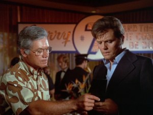 Duke, a Hawaiian man with grey and black hair wearing a light brown and dark brown aloha shirt, and Steve, a white man with brown hair wearing a dark blue suit and light blue shirt, discussing a case.