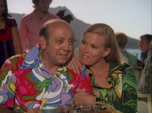 Heller is a middle aged, balding white man wearing a brightly colored Aloha shirt. Cindy is a beautiful blonde woman wearing a green aloha dress. She's smiling at him as he looks intently at someone off screen.