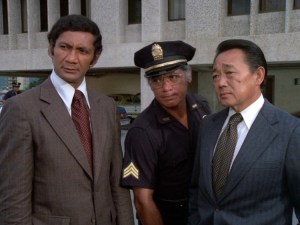 Al Harrington as Ben, a dark tan Pacific Islander man wearing a brown suit and maroon tie, Herman Wedemeyer as Duke, an older dark tan Pacific Island man wearing a dark blue police uniform and hat, and Kam Fong as Chin Ho Kelly, an tanned Asian man wearing a charcoal grey suit and brown tie. All three men look serious.