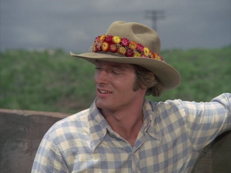 Perry King as Jay, a young white man with longish blond hair wearing a blue and white checked shirt and a brown cowboy hat with a red, orange, and yellow lei band.