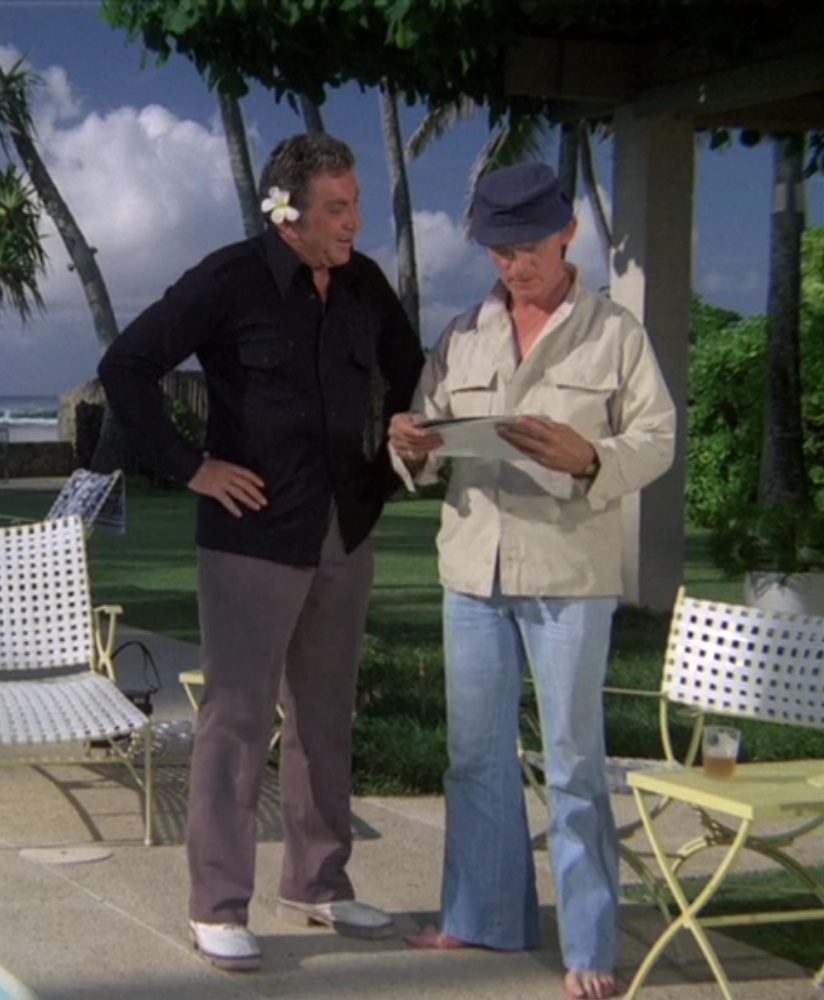 Cameron Mitchell, a middle aged white man with dark hair, wearing a dark blue shirt, grey pants, and white shoes with a white flower behind his ear, talking to Frank Gorshin, a middle aged white man wearing a blue hat, khaki shirt, and blue jeans, as he looks at some pictures. Two white chairs flank them and there's blue sky and palm trees behind them.