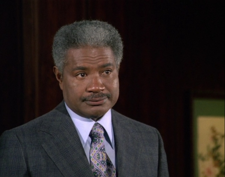Ossie Davis, a middle-aged Black man with salt and pepper hair and black mustache, wearing a dark grey patterned suit, purple patterned tied, and light purple shirt.