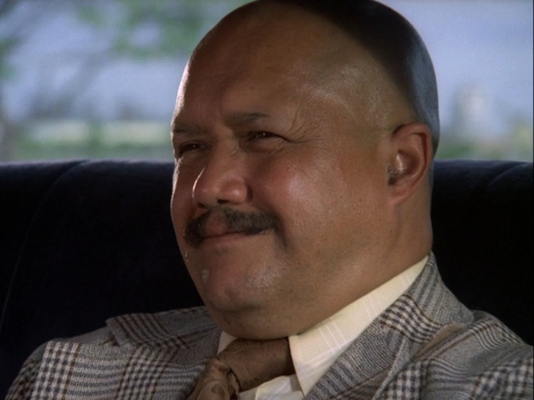 Wo Fat, a middle-aged Asian-appearing man, with a round, bald head and mustache, wearing a grey patterned suit and brown tie, sits smiling slightly in the backseat of a car.