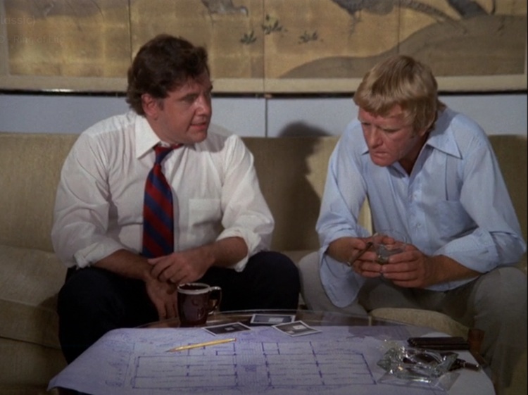 Clark, a middle aged, slightly heavyset white man with brown hair wearing a white buttoned down shirt and blue and red striped tie, and Nichols, a middle aged white man with blond hair wearing a light blue buttoned down shirt, sitting on a couch, looking at blueprints on a coffee table.
