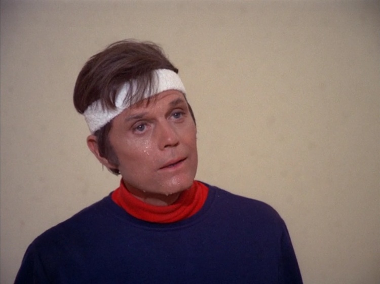 Jack Lord as Steve McGarret, a middle aged white man with brown hair that's being held off his forehead with a white sweatband. He's wearing a navy blue sweatshirt with a red collar. He's sweaty from playing handball.