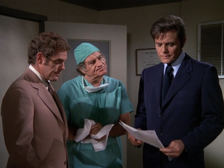 Danno, a young white man with curly dark blond hair wearing a dark tan suit, Doc Bergman, an older white man wearing teal scrubs, and Steve, a middle aged white man with brown hair and a dark blue suit at tie. They're looking at an autopsy report.