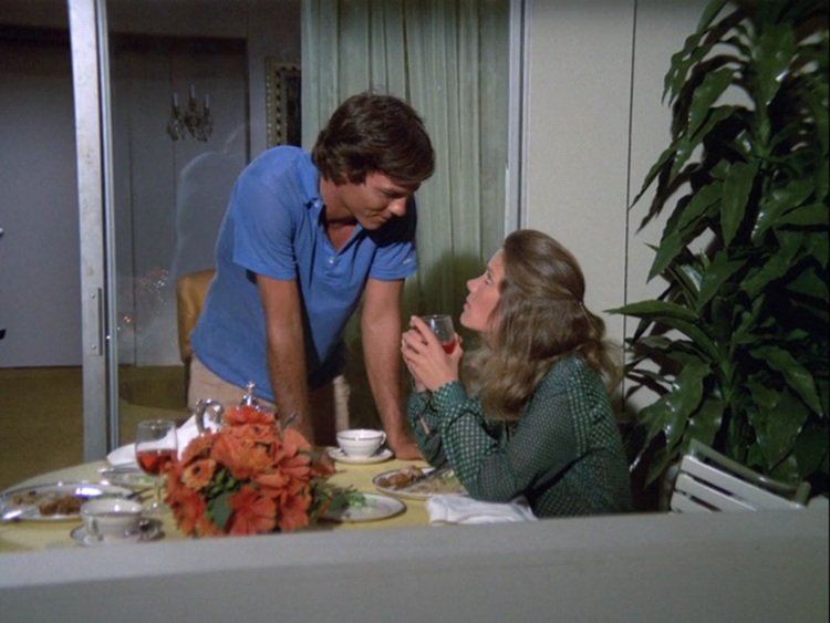 Richard Hatch, an extra tan young white man with dark hair wearing a blue polo shirt, leaning on a table looking down at Gretchen Corbett, a young white woman with long, dark blonde hair that's half pulled back. She's looking up at him and holding a glass.