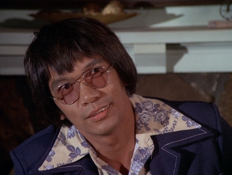 Rudolfo Aquino as Wanaka, a Pacific Islander man with longer, straight dark hair, wearing rose colored classes, a dark blue leisure suit, and a yellow, flower patterned shirt. The lapels are immaculate.