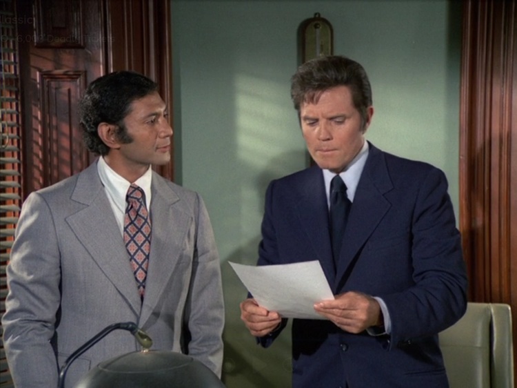 Al Harrington as Ben, a young Pacific Island man with black hair, excellent sideburns, wearing a grey suit with a pink and navy patterned tie and white shirt, and Jack Lord as Steve McGarrett, a middle aged white man with brown hair wearing a dark blue suit with matching tie and light blue shirt. Ben is looking at Steve and Steve is looking at a piece of paper.