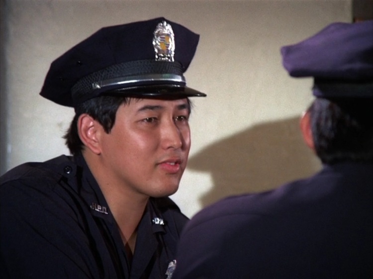 Dennis Chun as Officer Wade, a young Asian man wearing a dark blue police uniform and police hat.