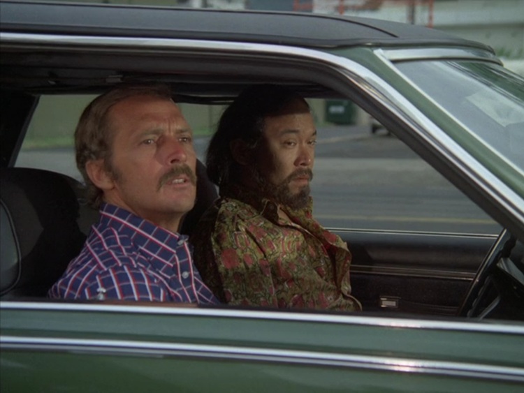 Jack Hogan, a middle aged white man with thinning blond hair and blond mustache wearing a blue, white, and red checked shirt, sitting in a car with this friend, a big Asian man with black hair, black beard, wearing a pink and green wildly patterned shirt.
