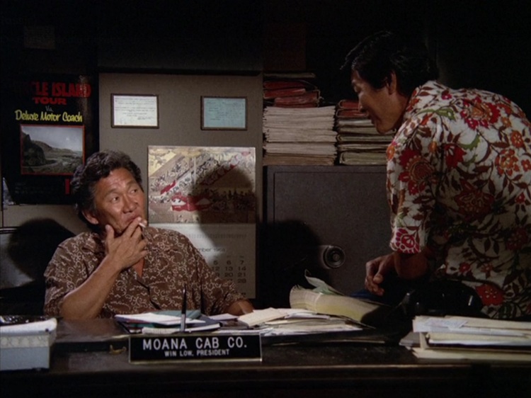 Kwan Hi Lim, an older Asian man with dark hair, wearing a brown and white patterned shirt and smoking a cigarette as he sits behind a desk, and Tommy Fujiwara, a middle aged Asian man with black hair wearing a red flower patterned shirt on a white background.