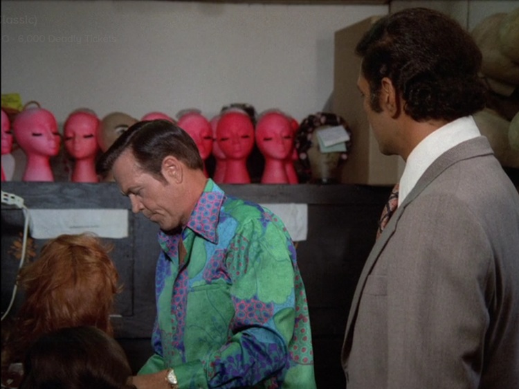 A hair stylist, a middle aged white man with brown hair and a green, pink, purple, and blue flower patterned shirt, and Ben, a young Pacific Island man with black hair wearing a grey suit. There's a row of pink mannequin heads behind them. It's slightly unsettling.